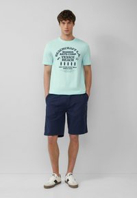 Lichtblauw graphic t-shirt met marineblauwe tekst, gecombineerd met donker marineblauwe shorts en witte sneakers met zwarte accenten. Eenvoudig ontwerp en pasvorm.