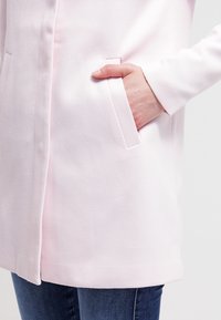 Ljusrosa, texturerad rock med en ensam bröstficka och en strukturerad design, kombinerad med mörkblå jeans.