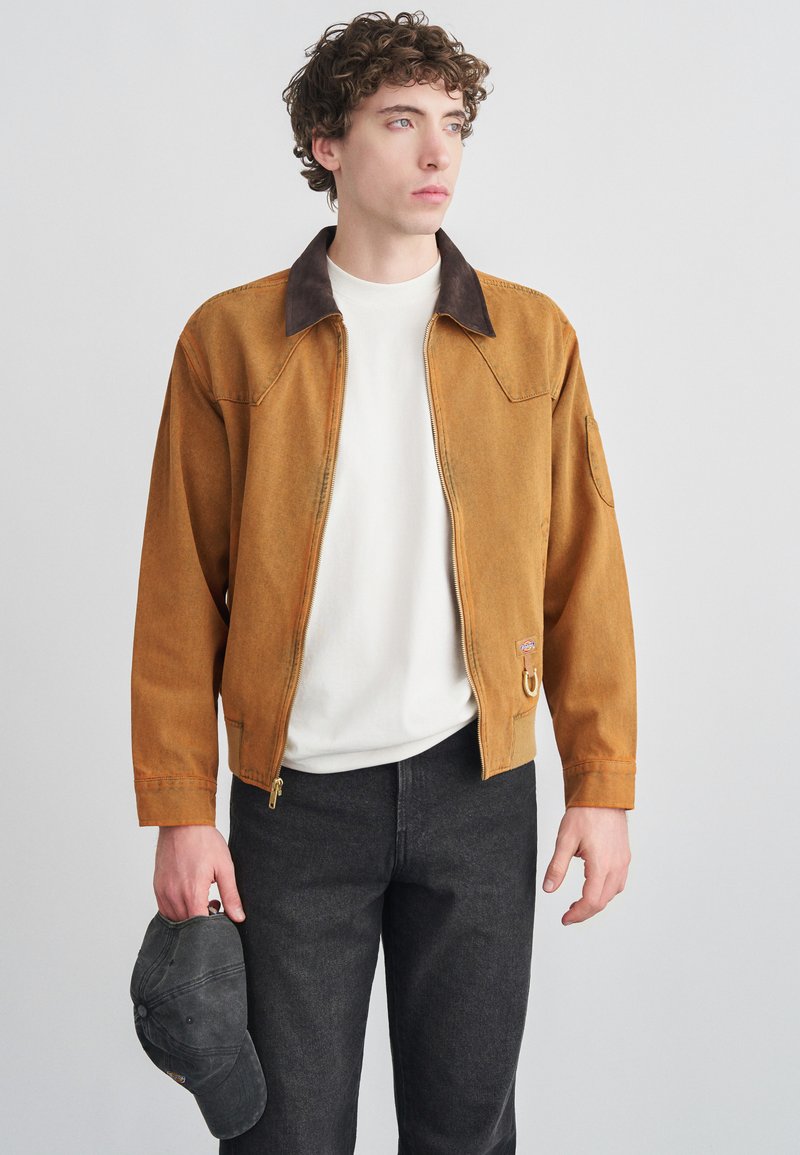 Καφέ μπουφάν με φερμουάρ και σκούρο γιακά, κατασκευασμένο από υφασμάτινη υφή. Συνδυάζεται με λευκό πουκάμισο και σκουρόχρωμα τζιν, κρατώντας ένα μαύρο καπέλο.