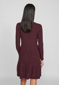 Maroonské šaty s ribovaným vzorem, dlouhými rukávy, vrstveným spodním lemem a přiléhavým tělem. Materiál vypadá měkký s texturovaným povrchem.