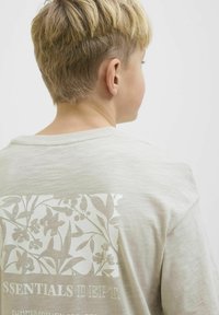 Lichtbeige T-shirt met korte mouwen en een textuur bloemenprint op de achterkant. Bevat de tekst "ESSENTIALS DEPT." onder het ontwerp.