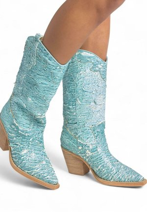 Botas de vaquero hechas de material turquesa texturizado con punta afilada y tacón de bloque de madera, que presentan un bordado intrincado y acentos blancos.