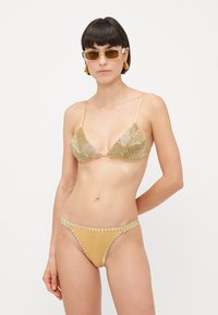 Vrouw in een met goud versierde bikini met driehoekige top en bijpassende broekjes, accessorized met gouden oorbellen en rechthoekige zonnebril.