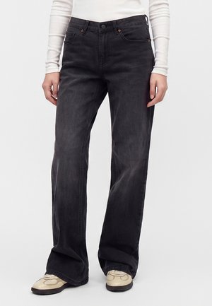 Person iført falmede sorte vidde jeans og hvide sneakers, med hænder afslappet langs siderne, mod ensfarvet lyst baggrund.