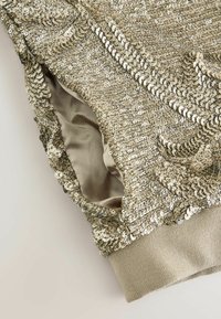 Sequined top in gold tones, featuring a textured surface with shiny sequins, soft beige lining, and a fitted cuff. Intricate detail design.