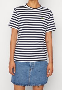 Person som bär en marinblå och vit randig T-shirt med en liten grön krokodil-logotyp, tillsammans med en blå denimkjol.