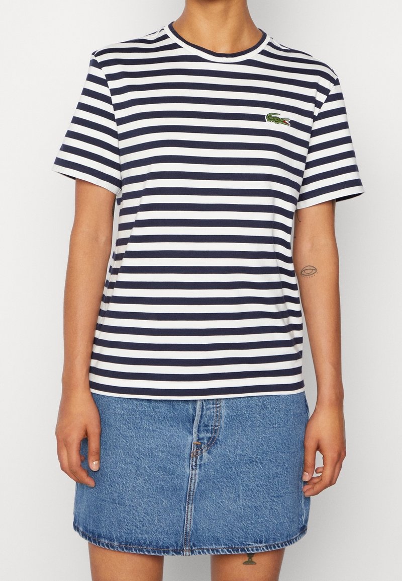 Person som bär en marinblå och vit randig T-shirt med en liten grön krokodil-logotyp, tillsammans med en blå denimkjol.