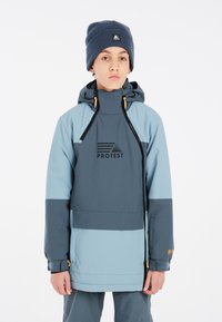 Veste imperméable bleue et grise avec un logo sur le devant. Dispose d'un col haut, d'une capuche réglable et de fermetures éclair contrastantes. Associée à un bonnet.