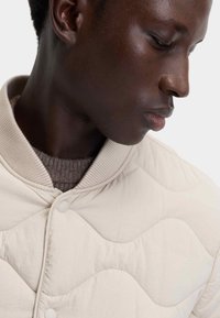 Beige quiltad jacka med ett texturerat vågmönster och ribbad krage, som har tryckknappar och en lager av stickat under.