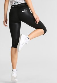 Kobieta w czarnych, krótkich legginsach Adidas autorstwa Stelli McCartney i białych sneakersach Adidas, podnosząca jedną nogę z tyłu w pozycji rozciągania.