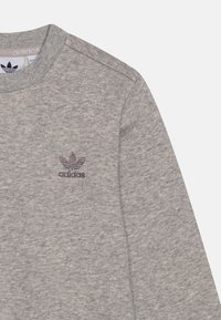 Sivi dukserica od mekog materijala ima okrugli izrez i izvezeni tamnoljubičasti Adidas logo na grudima.