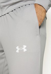 Gros plan d'une personne portant un pantalon de sport gris clair avec le logo Under Armour, main dans la poche, montrant la texture du tissu et la ceinture élastique.