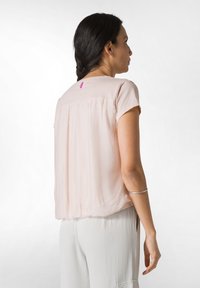 Ljusrosa skjorta gjord av mjukt tyg, med korta ärmar, lös passform och en rynkad design på baksidan. Inkluderar en liten lila etikett vid halsen.