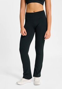 Leggings de fitness noirs et extensibles avec une taille haute. Texture lisse et design évasé à l'ourlet. Associés à des chaussures de sport blanches.