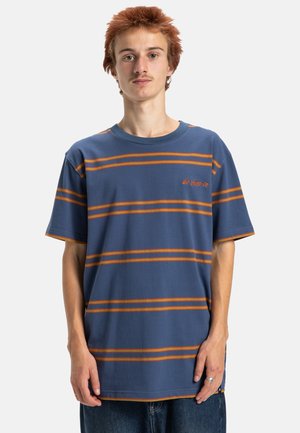 Młody mężczyzna z krótkimi czerwonymi włosami, ubrany w niebieską koszulkę z pomarańczowymi poziomymi paskami i niebieskie dżinsy, stojący na tle białym.