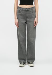 Osoba w luźnych szarych dżinsach, czarnym crop topie i czarno-białych trampkach, stojąca na tle jednolitego białego tła.