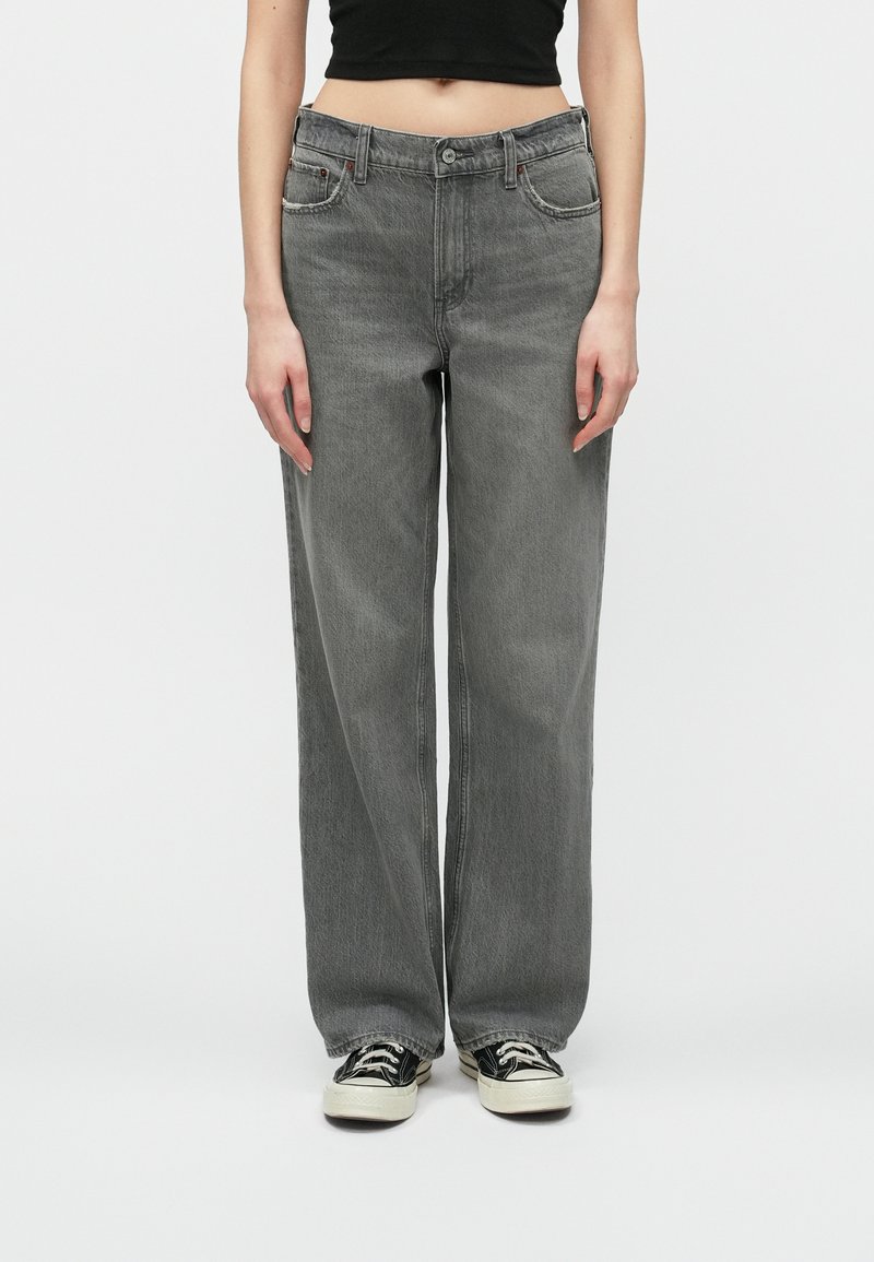 Osoba w luźnych szarych dżinsach, czarnym crop topie i czarno-białych trampkach, stojąca na tle jednolitego białego tła.