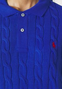 Polo shirt lavorato a maglia blu royal con design a treccia e colletto solido. Presenta tre bottoni e un piccolo logo rosso sul petto.