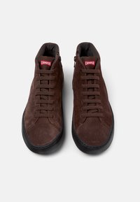 Bruine suede hoge sneakers met zwarte rubberen zolen, voorzien van veters aan de voorkant en zijritsen. Rode "CAMPER" logo op de tong.