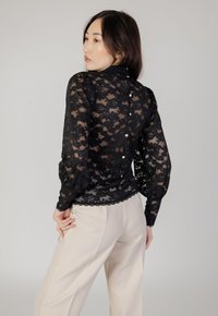 Schwarze Spitzenbluse mit langen Puffärmeln und einem gewellten Saum. Verfügt über Knopfdetails auf der Rückseite und eine halbdurchsichtige Textur.