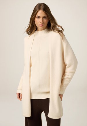 Cardigan lavorato a maglia color crema con apertura frontale, bordo a costine e maniche lunghe, abbinato a un top coordinato più chiaro e pantaloni marrone scuro.