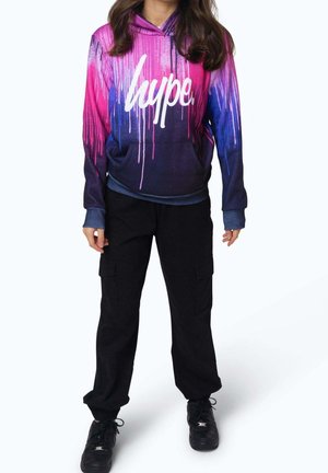 Hoodie met een verloop van roze en blauwe druppels, met witte tekst "lupe", gecombineerd met zwarte cargobroeken en zwarte sneakers.