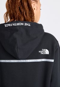 Svart hoodie tillverkad av bomull. Har en huva, vitt logotyp på baksidan och upprepande varumärkestext i grått över den nedre delen av ryggen.