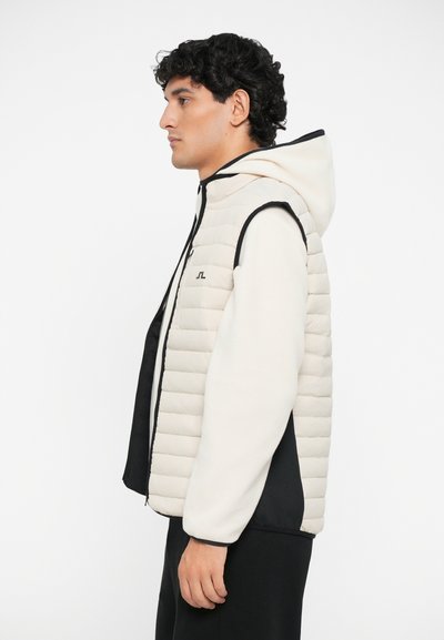 Puffervest i krem med svarte detaljer, med glidelås, hette og horisontalt quiltet mønster. Materialet ser mykt og lett ut.