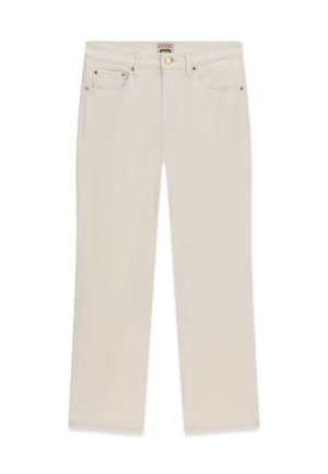 Jeune femme aux longs cheveux, portant un crop top noir et un pantalon blanc, debout sur des marches en pierre près d’un lierre feuillu et de l’eau.