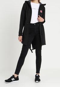 Giacca nera impermeabile con cappuccio grigio, dotata di chiusura con cordino e un'etichetta con logo. Abbinata a jeans skinny neri e sneakers nere.