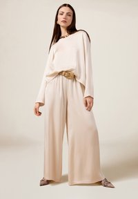 Beige satijnen top met wijde mouwen, bijpassende wijde broek en een opvallende gouden gespriemlint. Aangevuld met schoenen met een patroon van slangenprint.