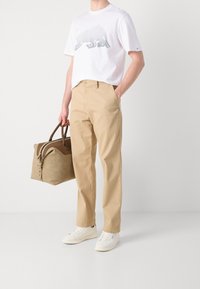 Valkoinen T-paita, jossa on pistekuvio, beige chino-housut, valkoiset lenkkitossut ja vaaleanruskea nahkakassi tummilla kahvoilla. Minimalistinen muotoilu ja rento tyyli.
