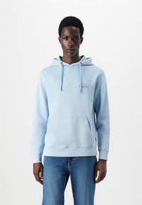 Gaišzilā kapuce, izgatavota no mīkstas auduma, ar kangaru kabatu, aukliņām un izšūtu logotipu uz krūts. Apvienota ar zilām džinsām.