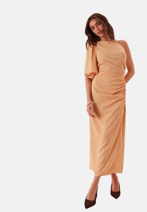 Femme portant une robe midi pêche asymétrique avec un volant froncé et une manche évasée, des talons noirs et un bracelet, debout devant un fond uni.