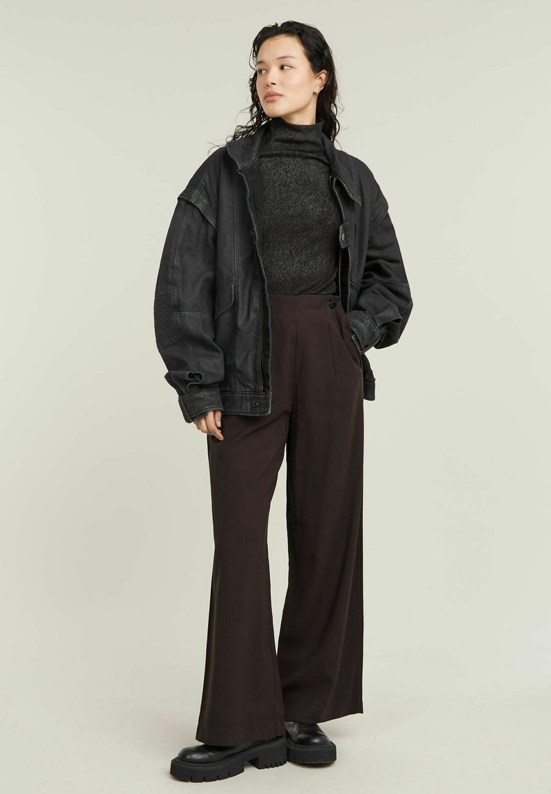 Kobieta w czarnej, oversize'owej kurtce, ciemnym swetrze z golfem, szerokich brązowych spodniach i masywnych czarnych butach, stoi z jedną ręką w kieszeni.