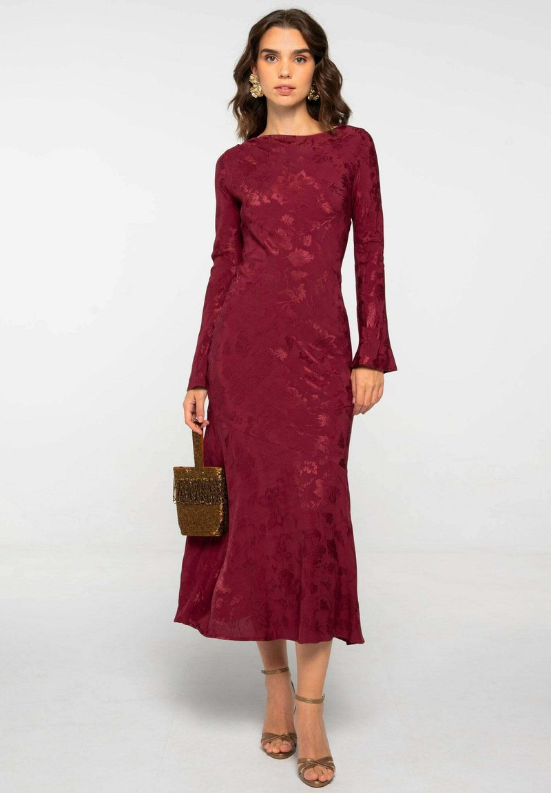 Vestido burdeos de manga larga con textura floral, cuerpo entallado, dobladillo acampanado, acompañado de un pequeño bolso de mano dorado y tacones beige con tiras.