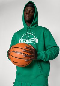 Zöld Celtics kapucnis pulóvert viselő férfi, kezében narancssárga „playground” márkájú kosárlabdával, a kamerába néz egy egyszínű háttér előtt.