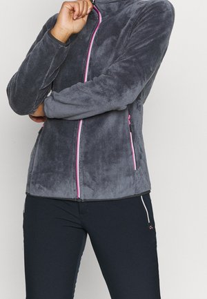 Person wearing a dark gray fleece jacket with pink zipper details and dark pants, with one arm crossed and the other hand touching their face.