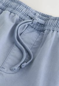 Pantalon à taille élastique bleu clair avec un cordon de serrage noué et des coutures de poche avant visibles sur un tissu lisse.