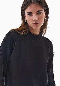 Czarny sweatshirt z wysokim kołnierzem i długimi rękawami. Miękka tekstura, luźny krój i minimalne detale szwów. Okrągły, prążkowany kołnierz.