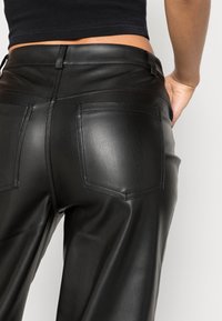 Pantalon en cuir noir avec une texture lisse, doté de deux poches arrière et d'une coupe ajustée. La taille se situe juste en dessous de la taille naturelle.