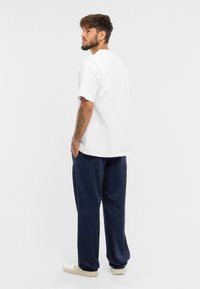 Biele tričko s krátkym rukávom a okrúhlym výstrihom, kombinované s námořníckymi teplákmi a bielymi teniskami. Viditeľné tetovania na ramene.