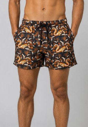 Uomo che indossa pantaloncini da bagno neri e marroni con motivi di serpente arancioni e coulisse nera, mani nelle tasche, su sfondo grigio.