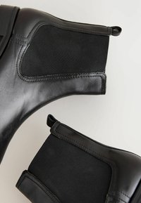Las botines negros presentan un exterior de cuero elegante y paneles laterales elásticos, mostrando un diseño moderno sobre un fondo neutro.
