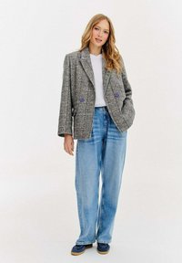 Žena s dlouhými blond vlasy v šedém kostkovaném saku, bílé košili, modrých džínách s volnými nohavicemi a námořnických teniskách, stojící na bílém pozadí.