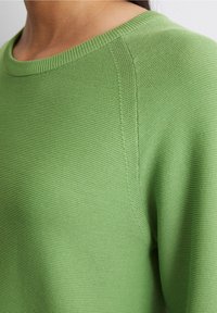 Zbliżenie na osobę ubraną w jasnobrązowy sweter o teksturowanym splocie z okrągłym dekoltem, ukazujące detale na ramionach i szyi.