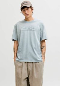 Hellblaues Baumwoll-T-Shirt mit kurzen Ärmeln, das ein aufgedrucktes Logo in dunklerem Blau zeigt. Kombiniert mit beigen, plissierten Hosen.