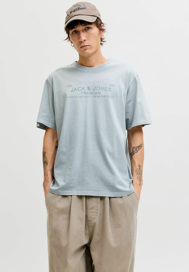 Hellblaues Baumwoll-T-Shirt mit kurzen Ärmeln, das ein aufgedrucktes Logo in dunklerem Blau zeigt. Kombiniert mit beigen, plissierten Hosen.