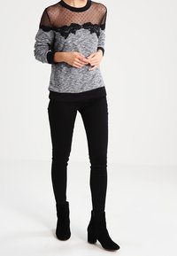 Suéter gris con un yugo de malla negra transparente con puntos y adornos de encaje, combinado con pantalones skinny negros y botas de tobillo negras.