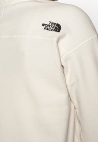 Ljus krämfärgad fleece-tröja med The North Face-logga i svart, med synliga sömmar och en avslappnad passform på axeln.